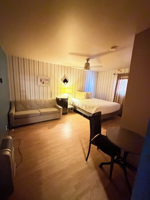 Hotelkamer met een bed, bureau, stoel, bank, nachtkastje, lamp, plafondventilator en raam met gordijnen.