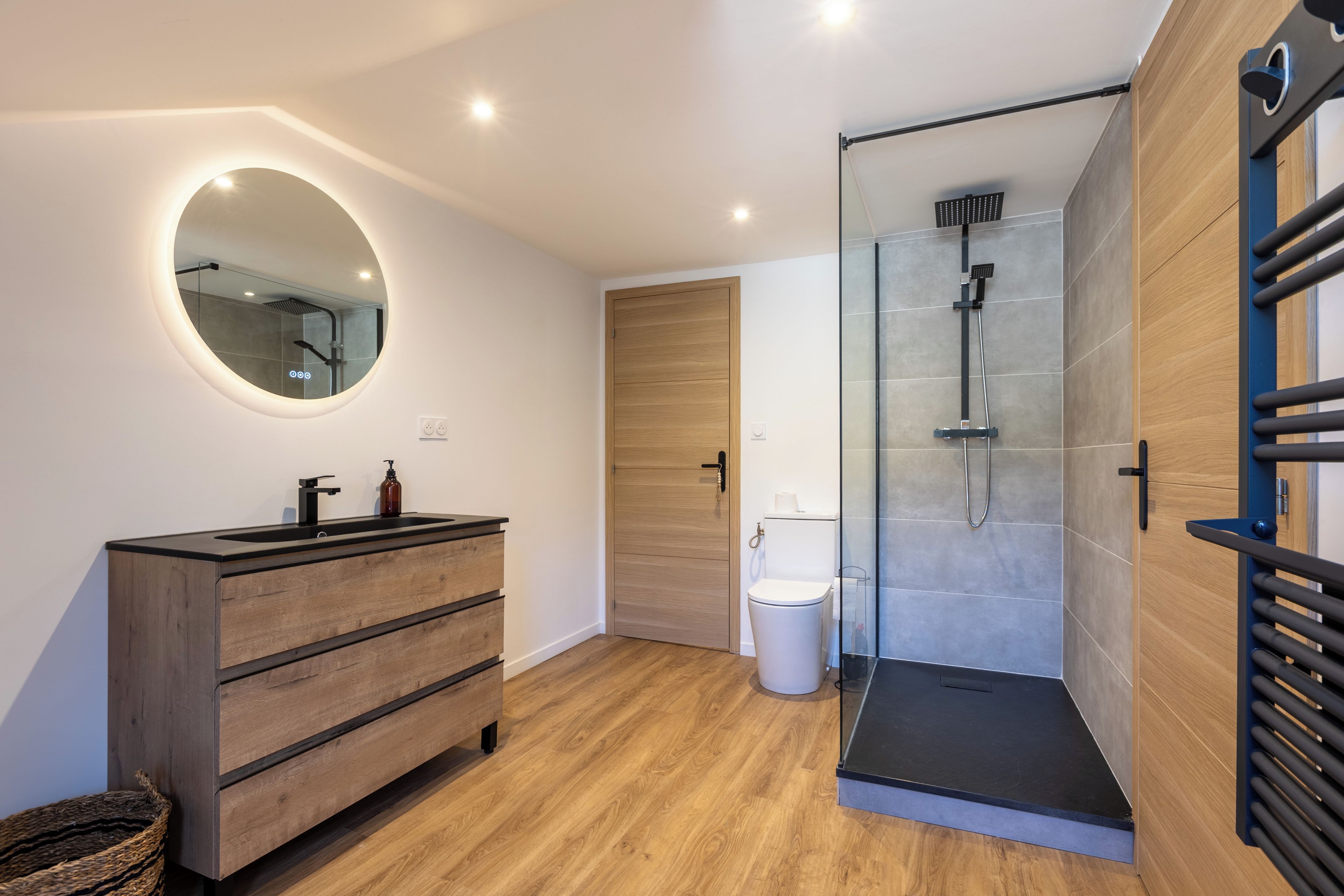 A modern bathroom with a wooden vanity, a round mirror, a glass shower enclosure, and a toilet.