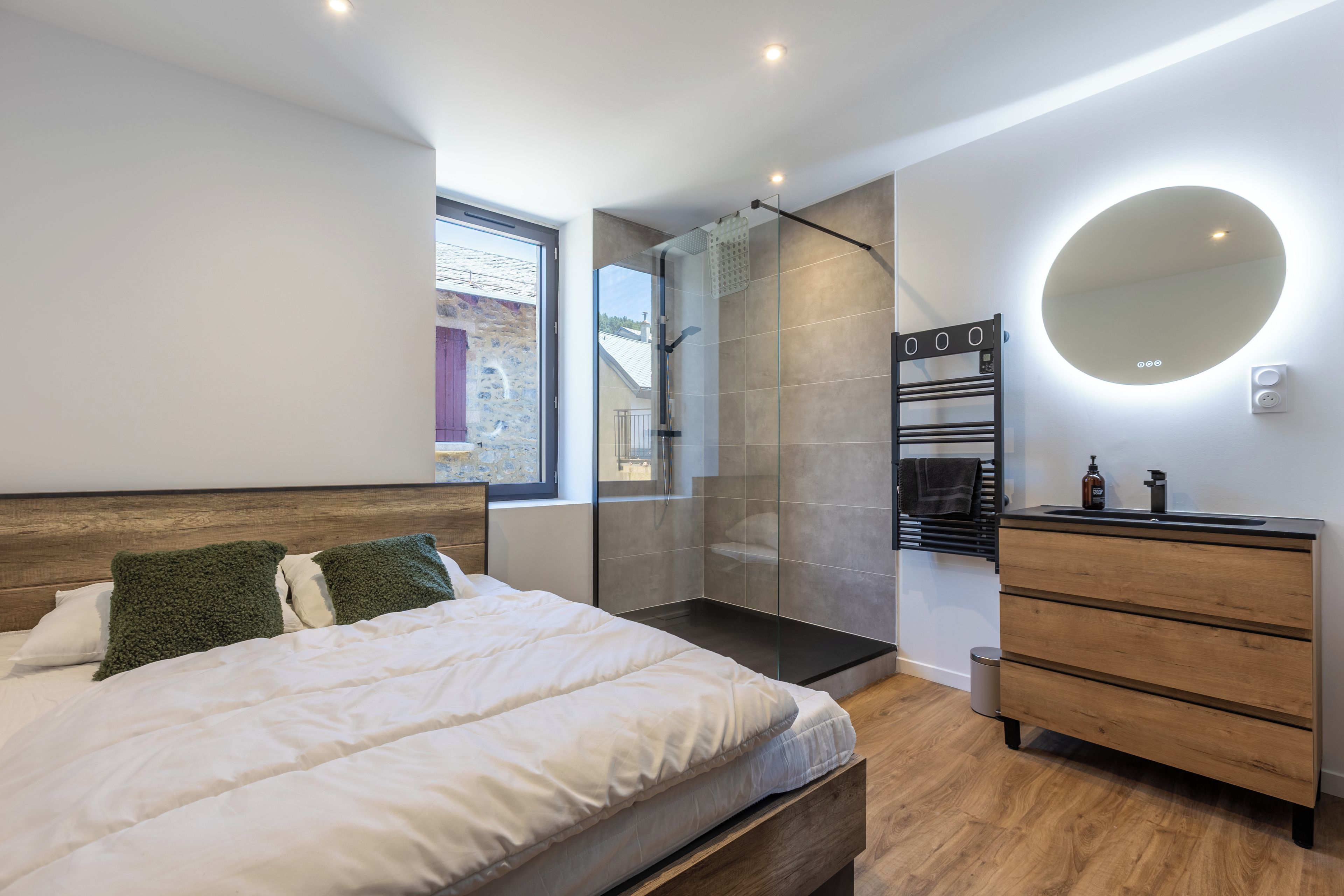 Ein modernes Schlafzimmer mit einem Bett, einer Holzkommode, einem Spiegel und einer Duschkabine aus Glas.