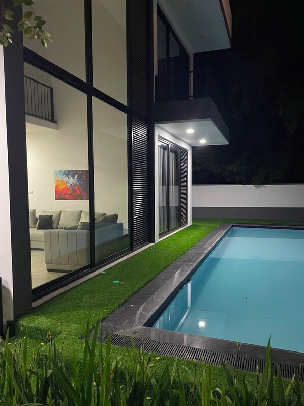 Una casa moderna con piscina, un ventanal que da a una sala de estar y una zona ajardinada.