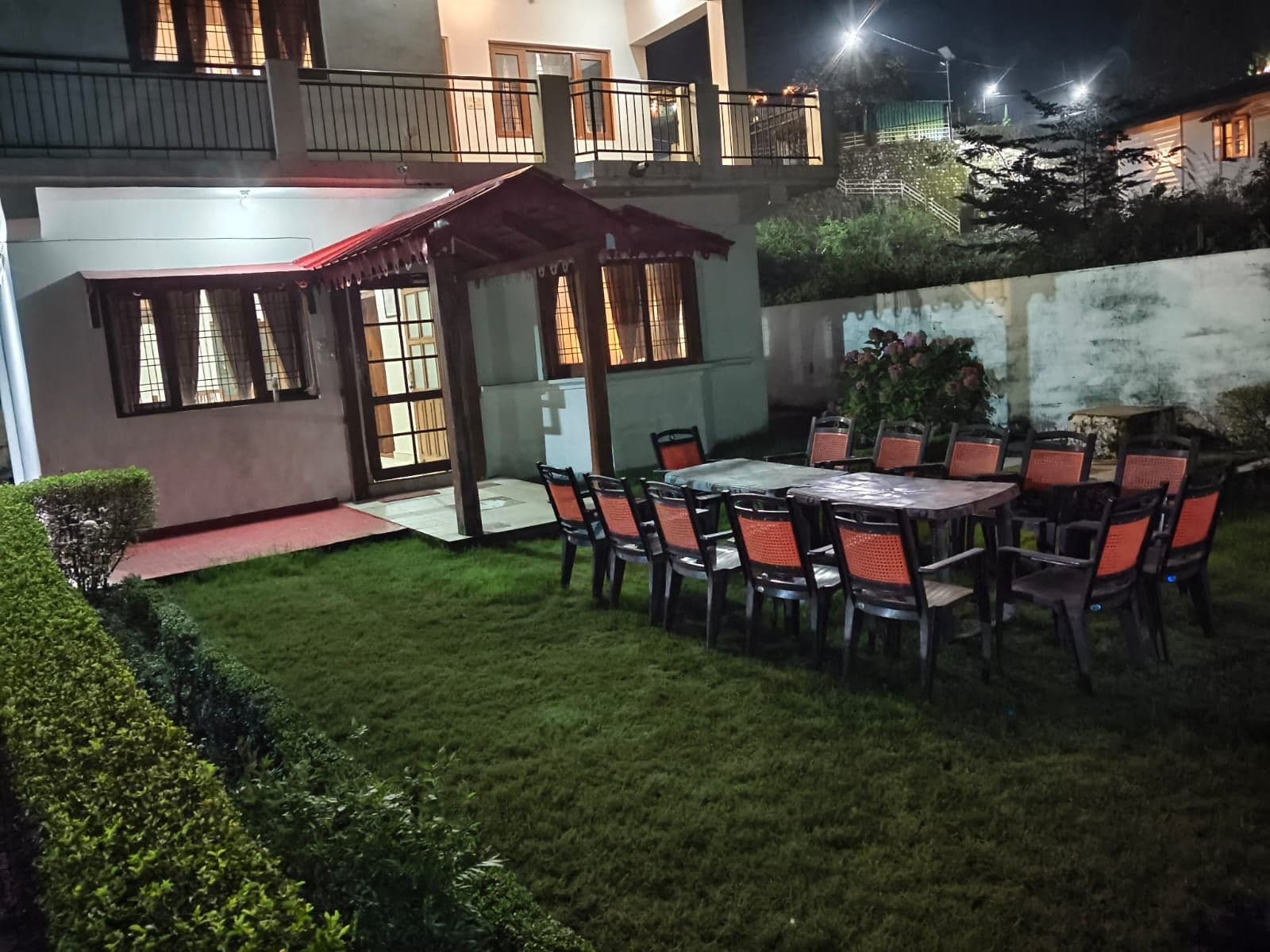 A well-lit outdoor patio area with a dining table and chairs, surrounded by a neatly trimmed lawn and a small garden.