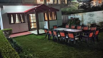 A well-lit outdoor patio area with a dining table and chairs, surrounded by a neatly trimmed lawn and a small garden.