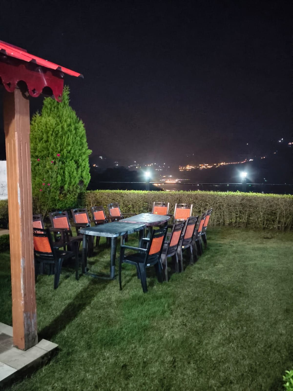 An outdoor seating area with a table and chairs, a red roof structure, and a hedge in the background.