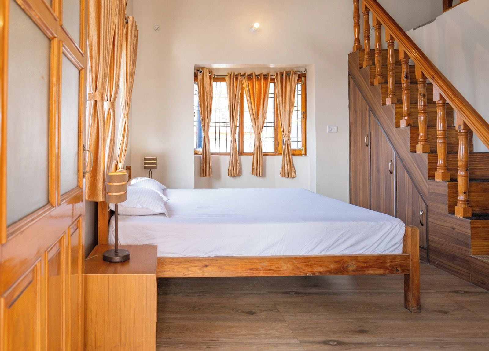 A bedroom with a wooden bed, a nightstand, a lamp, a window with curtains, and a wooden staircase.