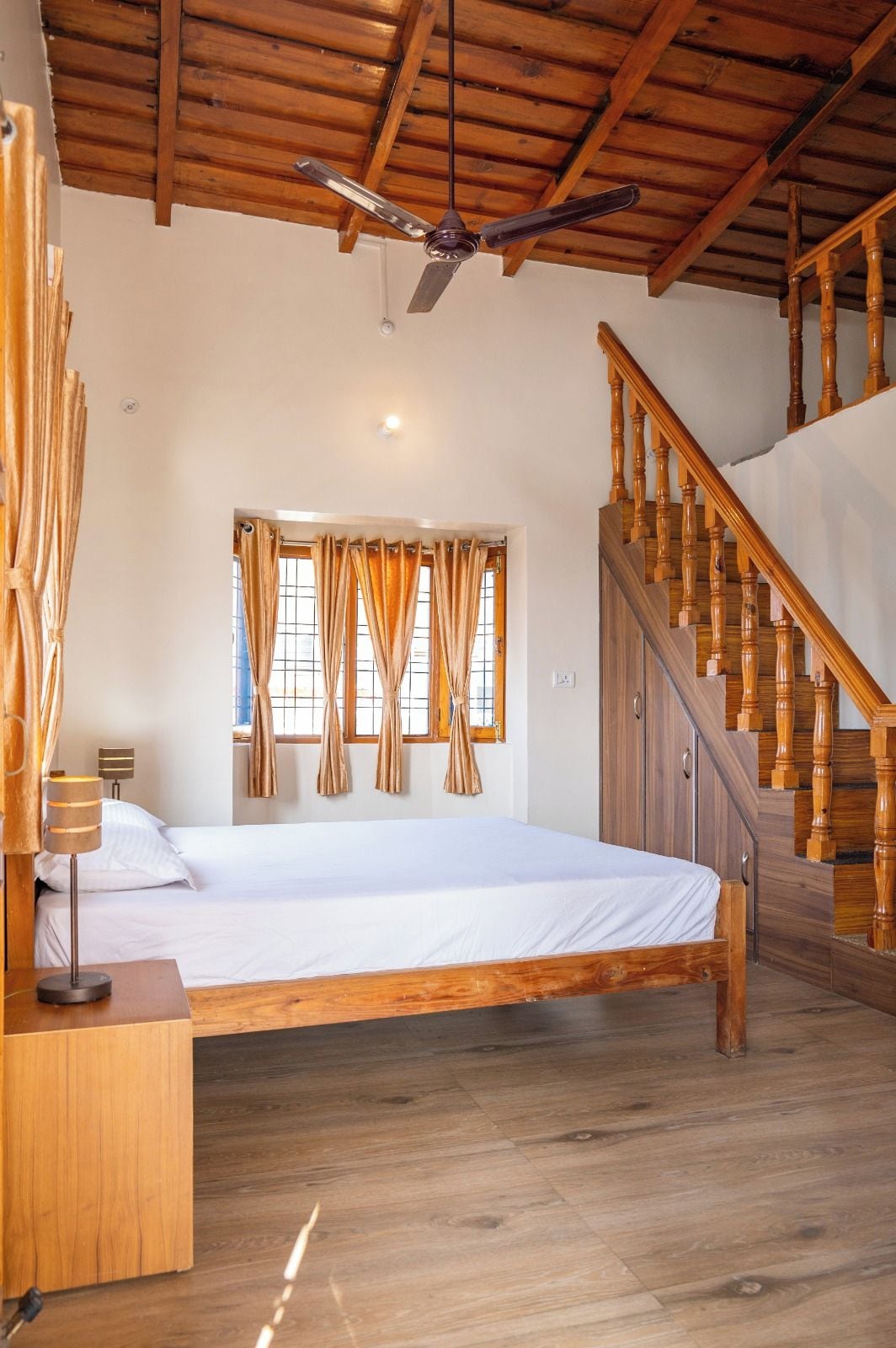 A bedroom with a wooden bed, a ceiling fan, a staircase, and a window with curtains.