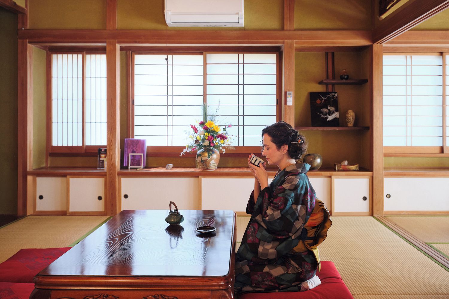 Ein traditioneller japanischer Raum mit Tatami-Böden, einem niedrigen Holztisch und einer Person, die auf einem roten Kissen sitzt.