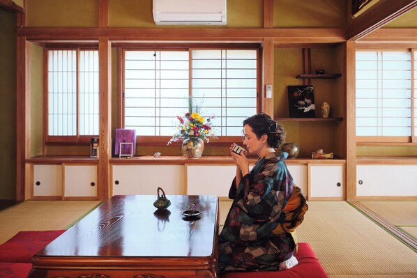 A traditional Japanese room with tatami flooring, a low wooden table, and a person sitting on a red cushion.