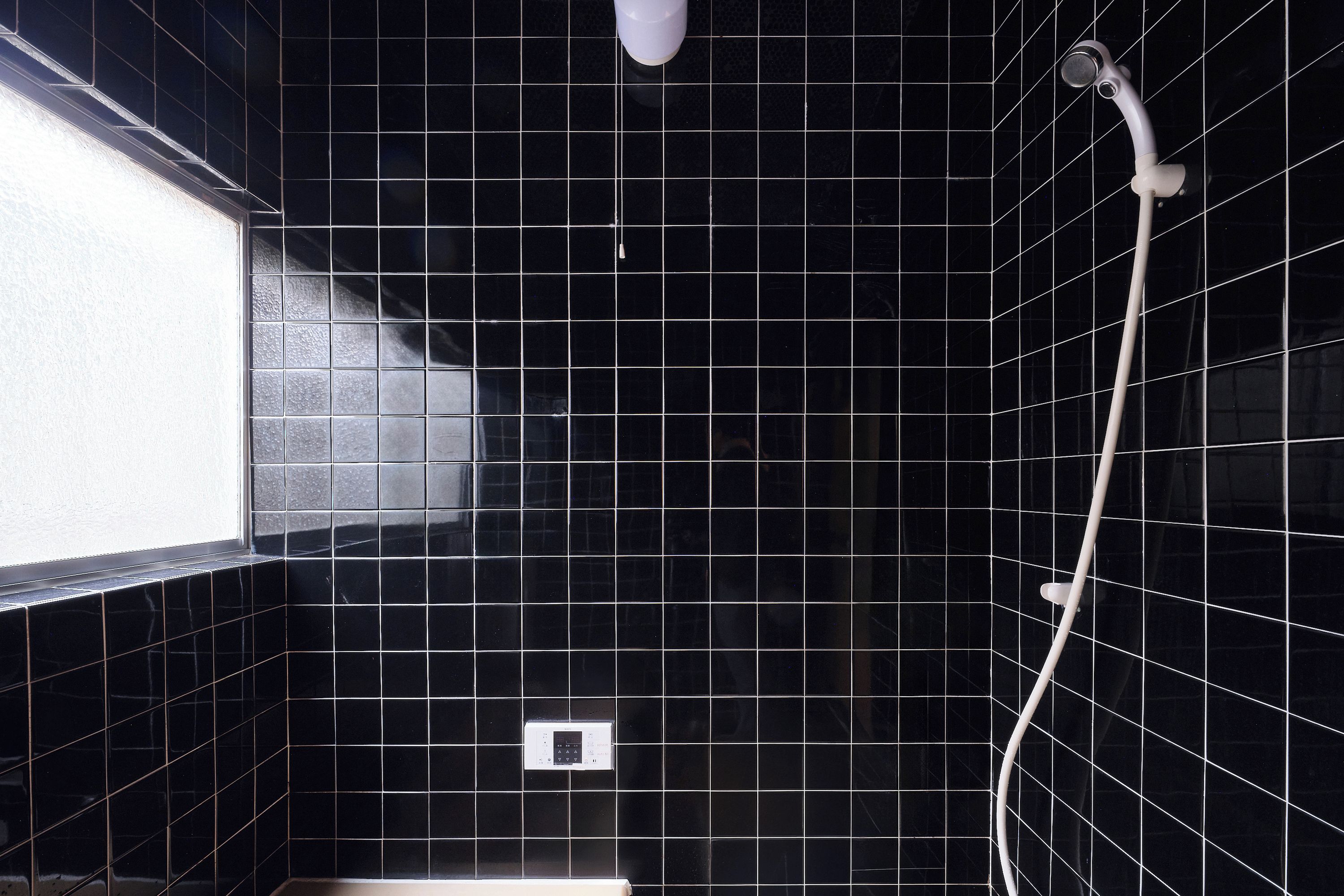 A bathroom with black tiled walls, a window on the left, and a shower with a handheld showerhead.