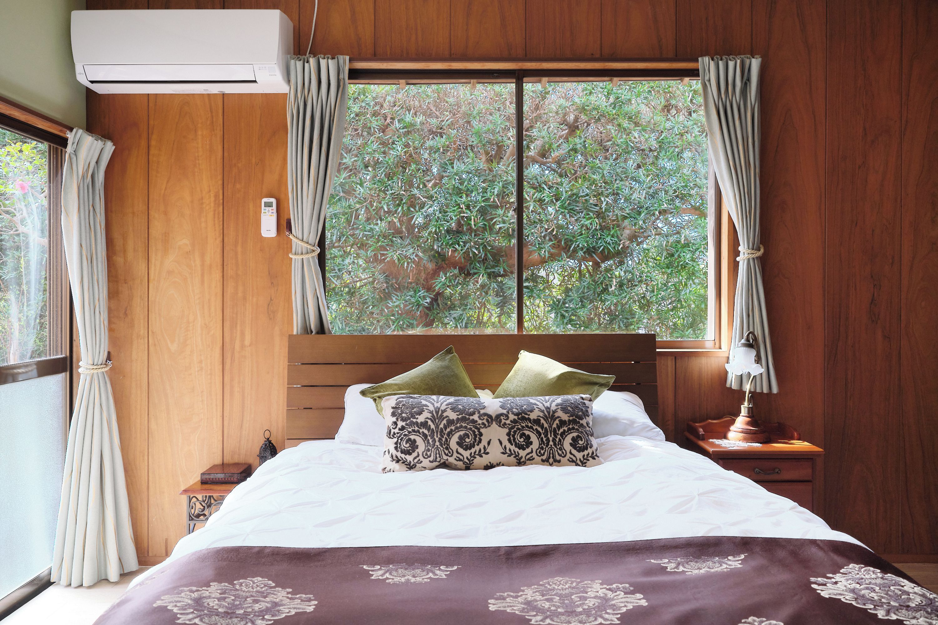A bedroom with a wooden headboard, a bed with white bedding and patterned pillows, a bedside table with a lamp, and a window with curtains.