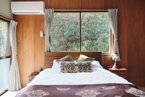A bedroom with a wooden headboard, a bed with white bedding and patterned pillows, a bedside table with a lamp, and a window with curtains.