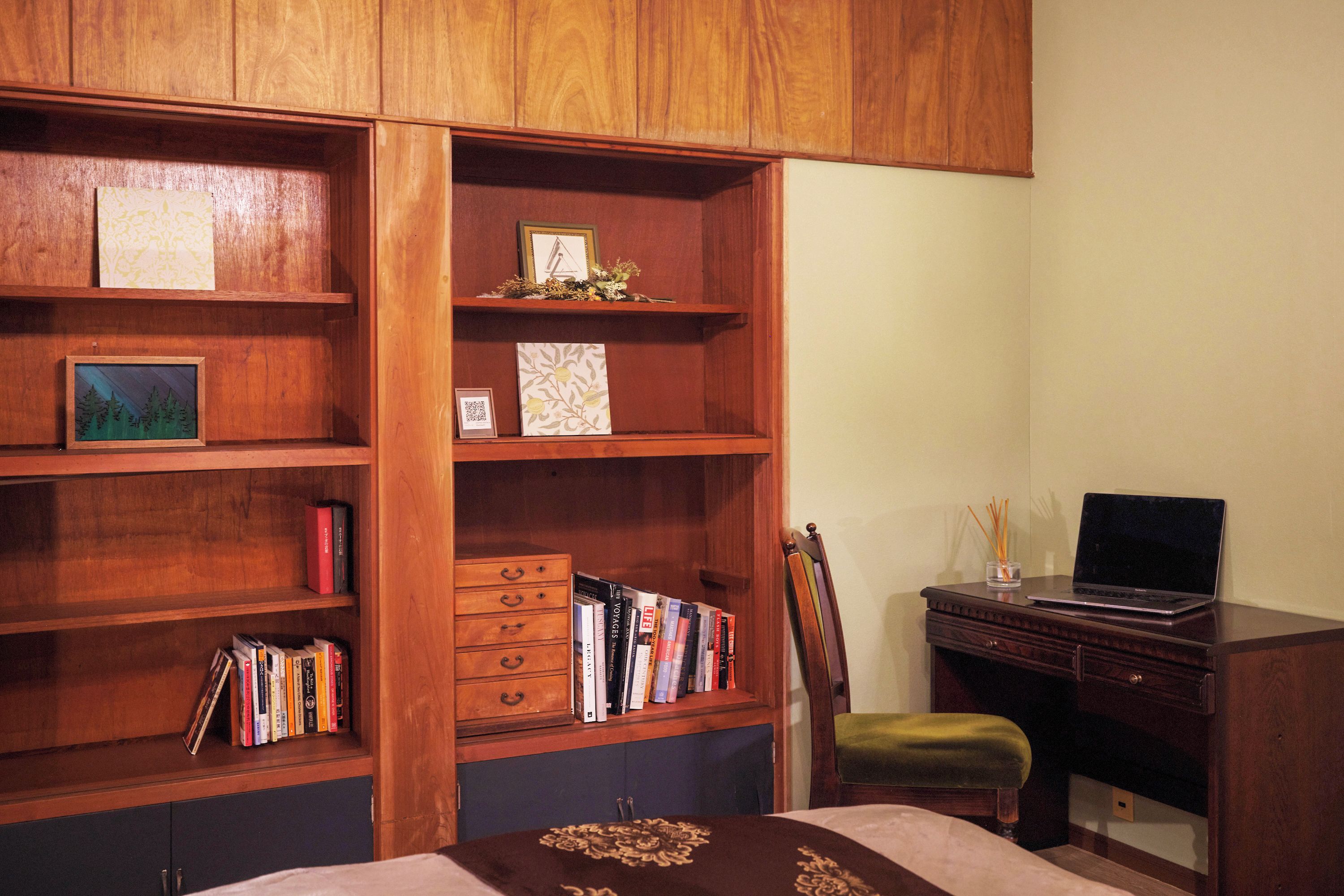 A room with a wooden bookshelf containing books and framed pictures, a desk with a laptop, and a chair.