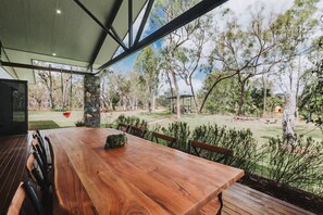 An outdoor dining area with a wooden table and chairs, surrounded by a stone pillar and a view of a grassy yard with trees. - The Riverstone | Luxury Bush Retreat (Millstream)
