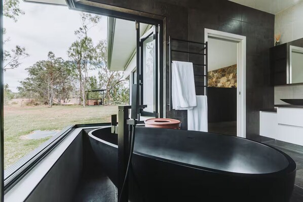 A modern bathroom with a large black bathtub, a glass shower enclosure, and a view of the outdoors.