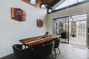 A modern dining area with a wooden table, black chairs, and a glass door leading to an outdoor patio. - The Riverstone | Luxury Bush Retreat (Millstream)