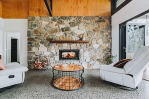 A cozy living room with a stone fireplace, a round coffee table, and two white armchairs. - The Riverstone | Luxury Bush Retreat (Millstream)