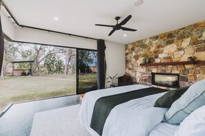 A bedroom with a large bed, a stone wall, a fireplace, and a view of the outdoors. - The Riverstone | Luxury Bush Retreat (Millstream)