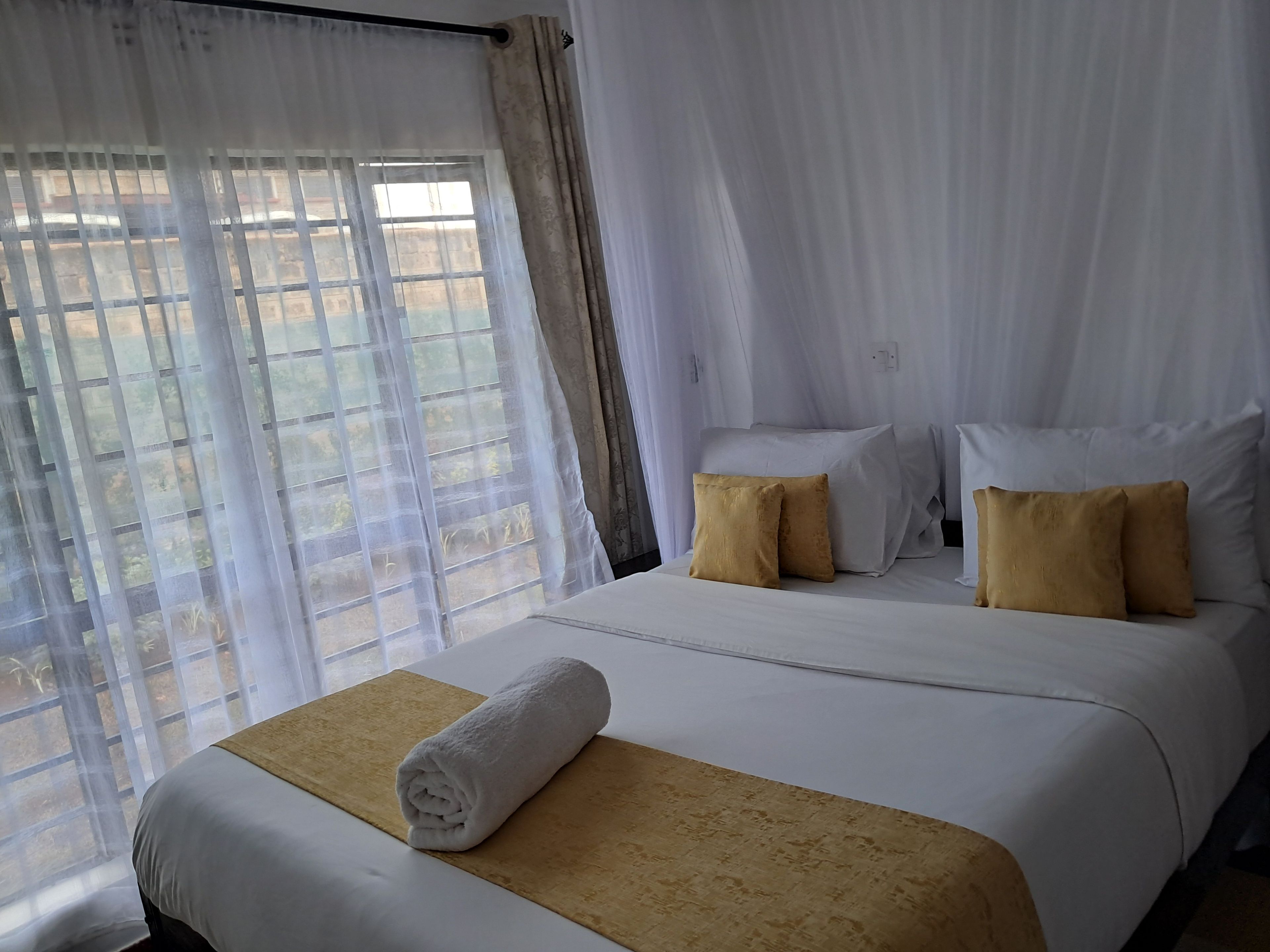 A hotel room with a bed, two pillows, a rolled towel, and a window with curtains.