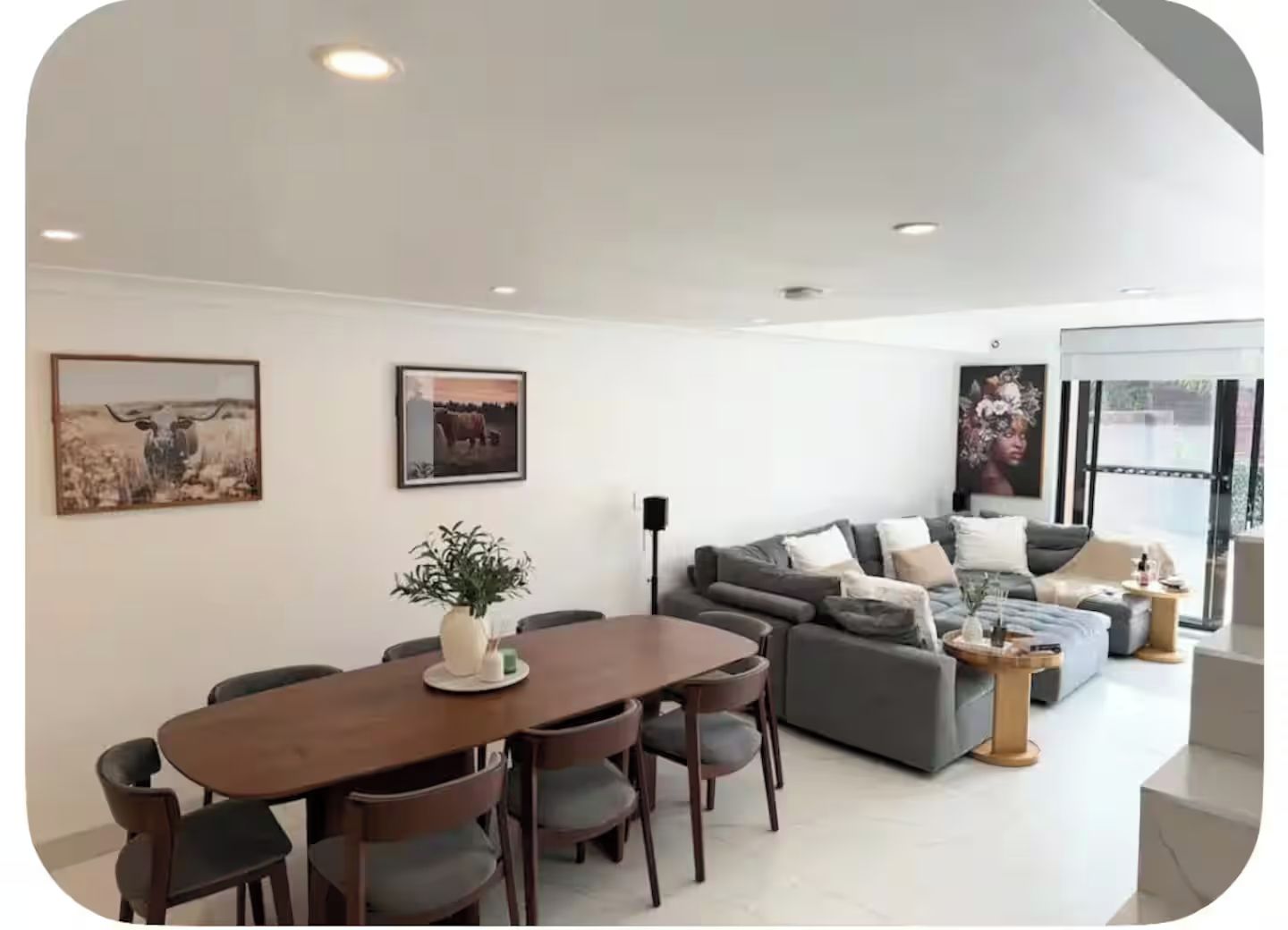A modern living room with a dining area, featuring a sofa set, a dining table with chairs, and two wall-mounted paintings.