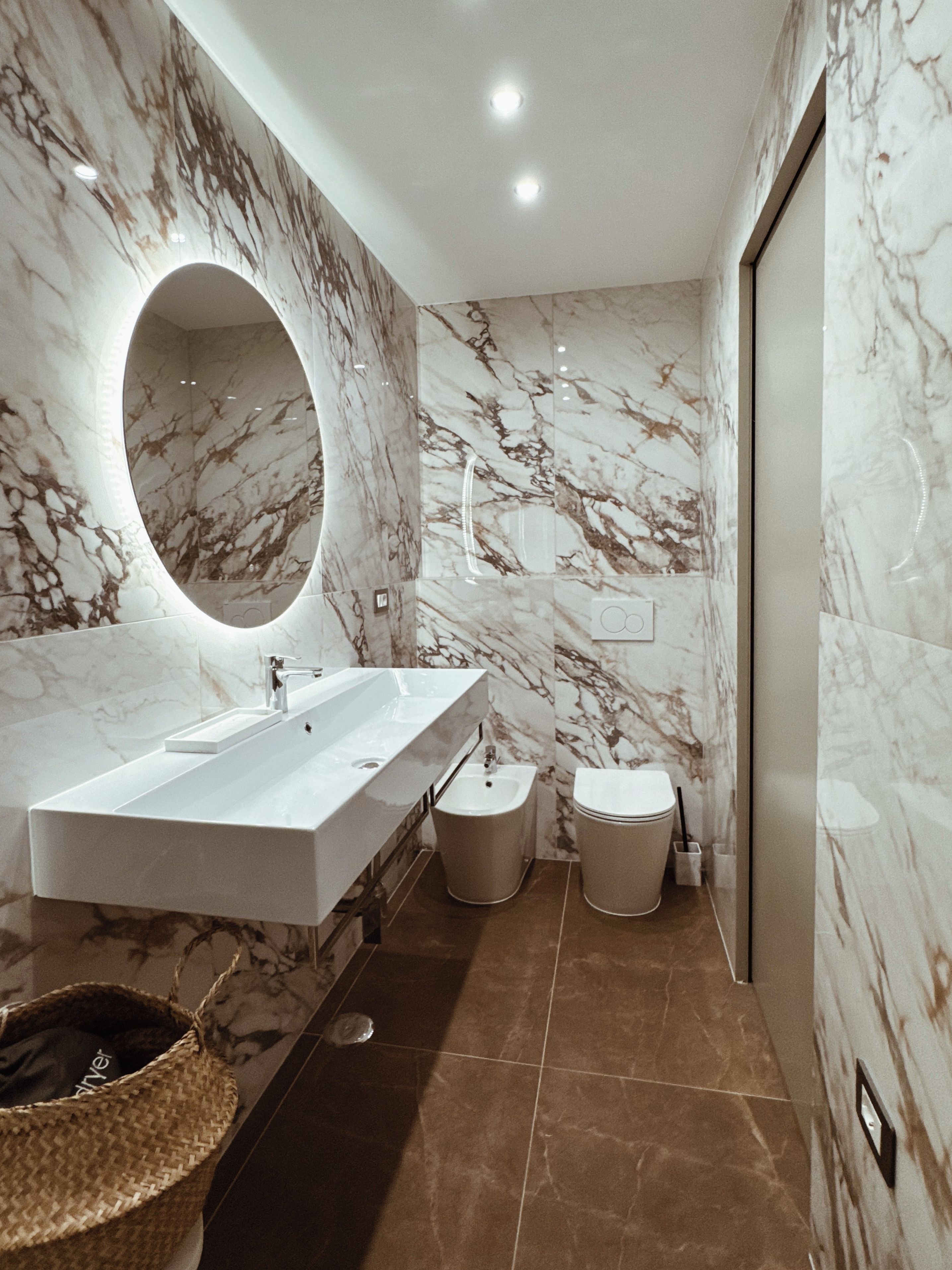 Un baño moderno con un espejo ovalado grande, un lavamanos rectangular, un bidé y un inodoro.
