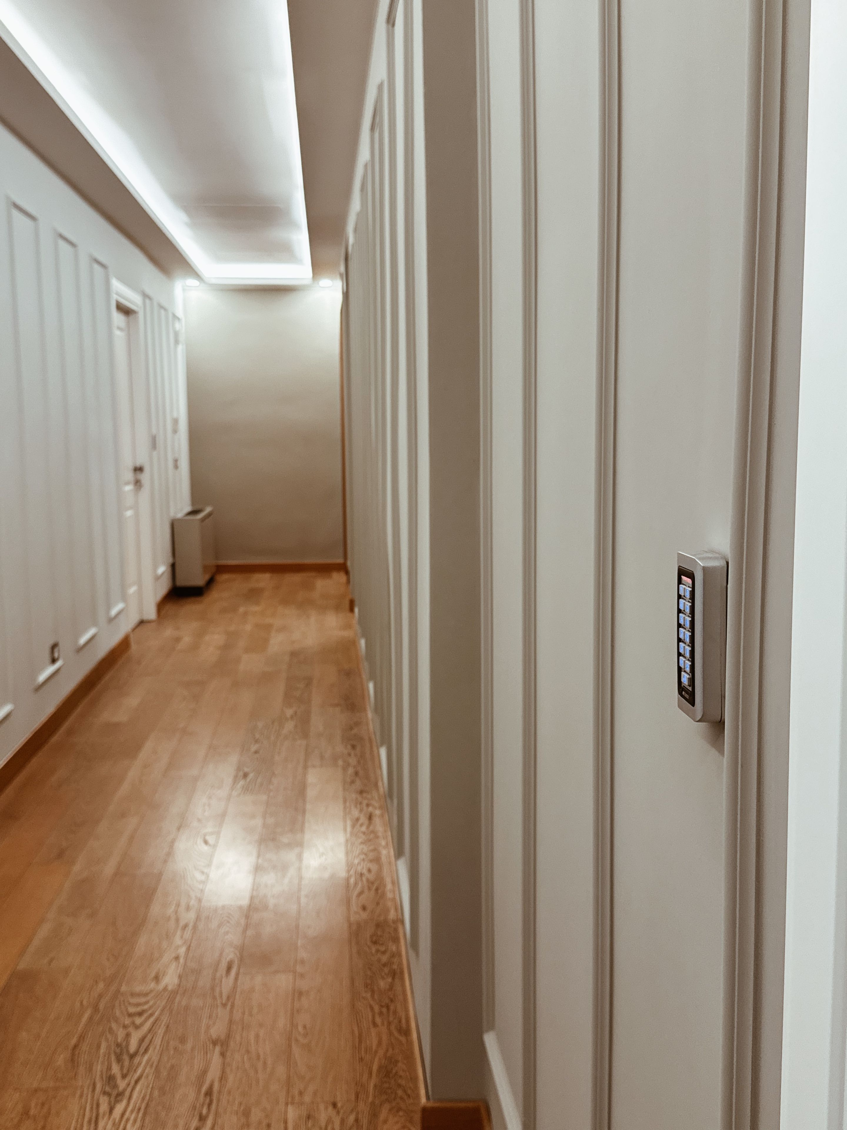 Un pasillo con piso de madera, paredes blancas y una puerta con cerradura electrónica.