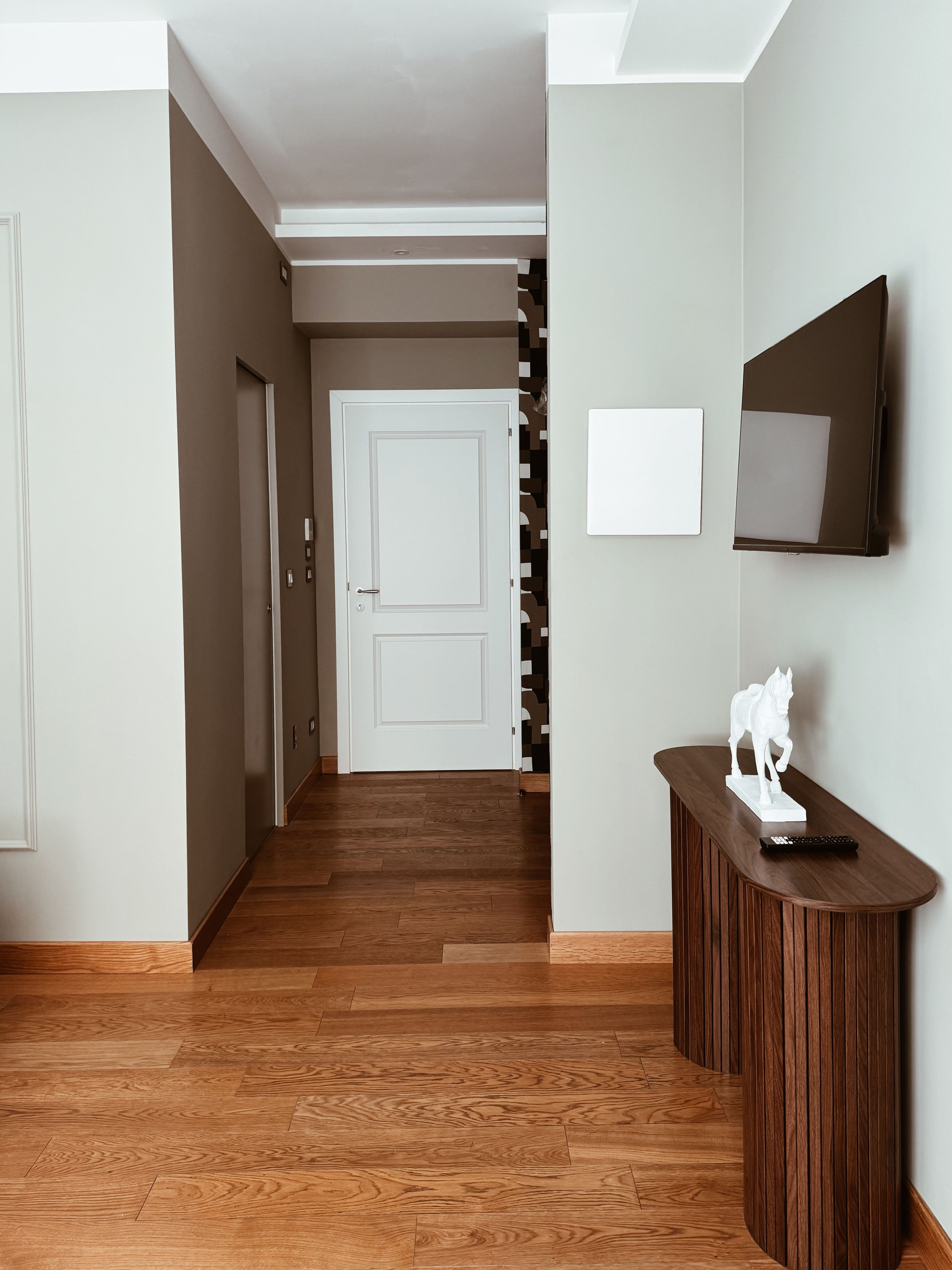 Un pasillo con piso de madera, una puerta blanca, un televisor de pared y una pequeña estatua de un caballo blanco sobre una consola de madera.