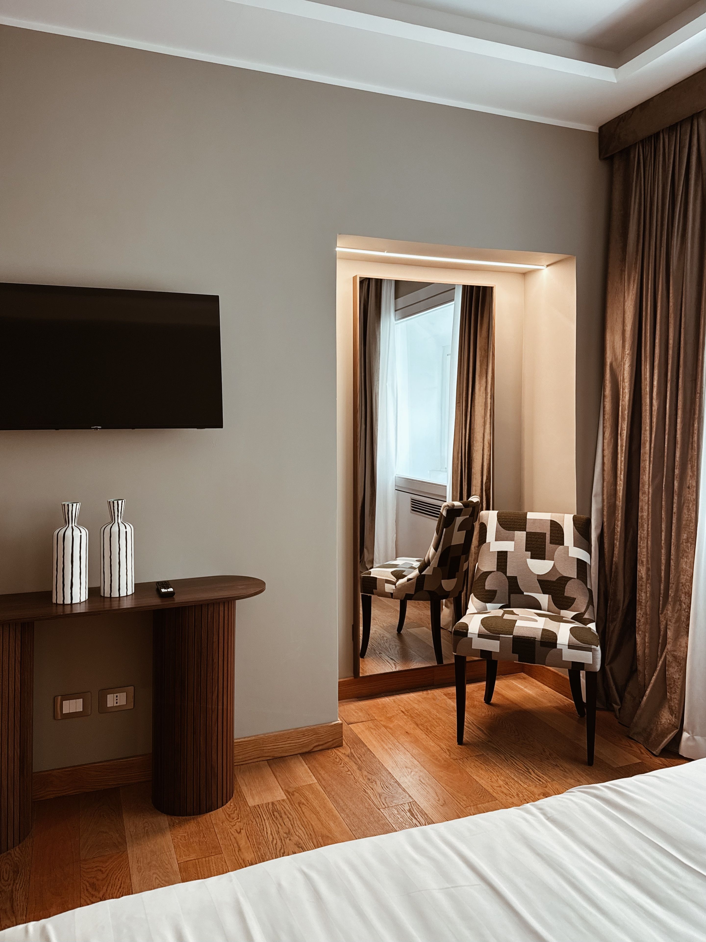 Habitación de hotel con cama, televisor montado en la pared, mesita de madera con dos jarrones, sillón estampado y un espejo que refleja la habitación.