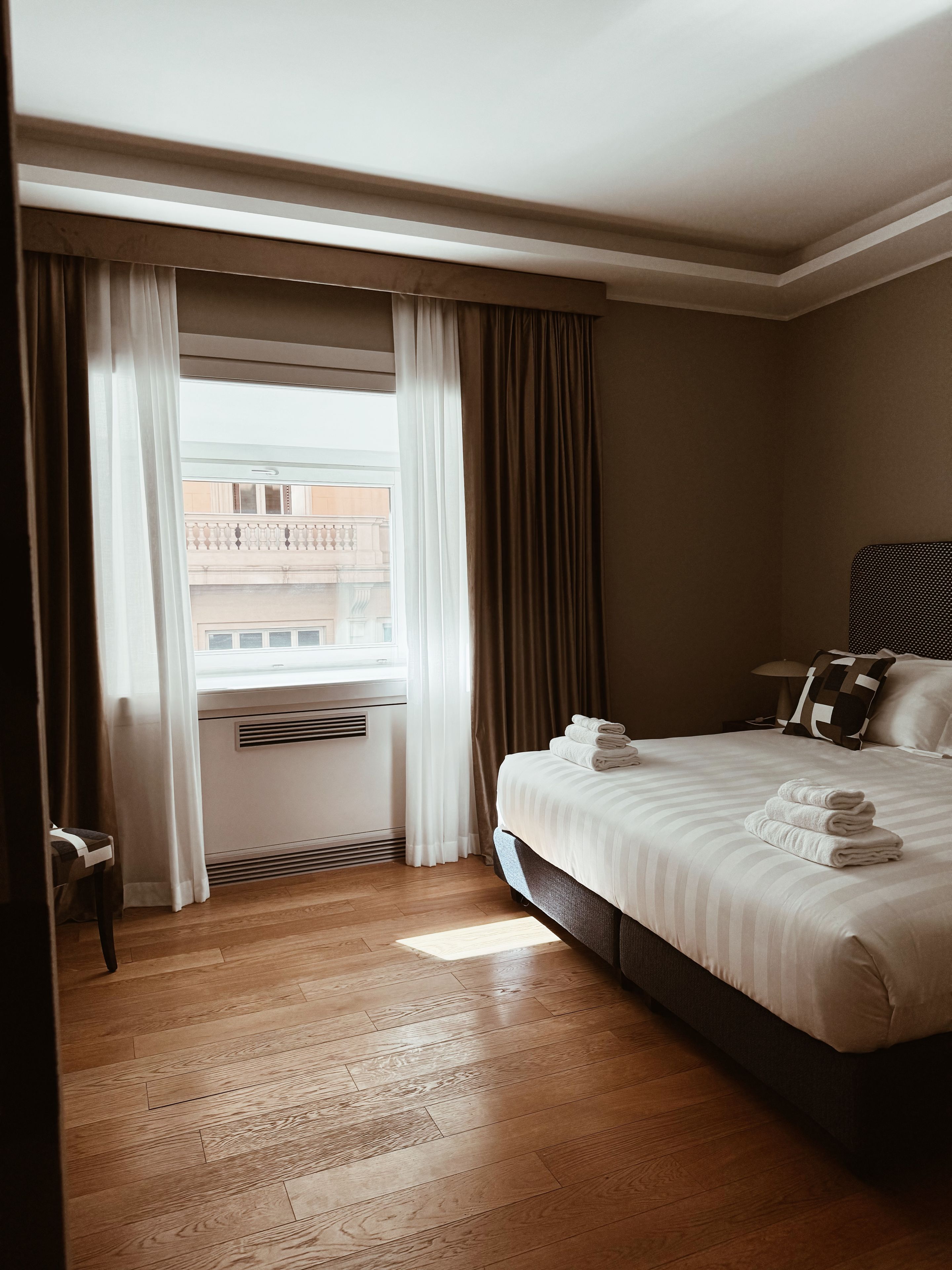 Habitación de hotel con una cama grande, ventana con cortinas, piso de madera y vista a edificios.