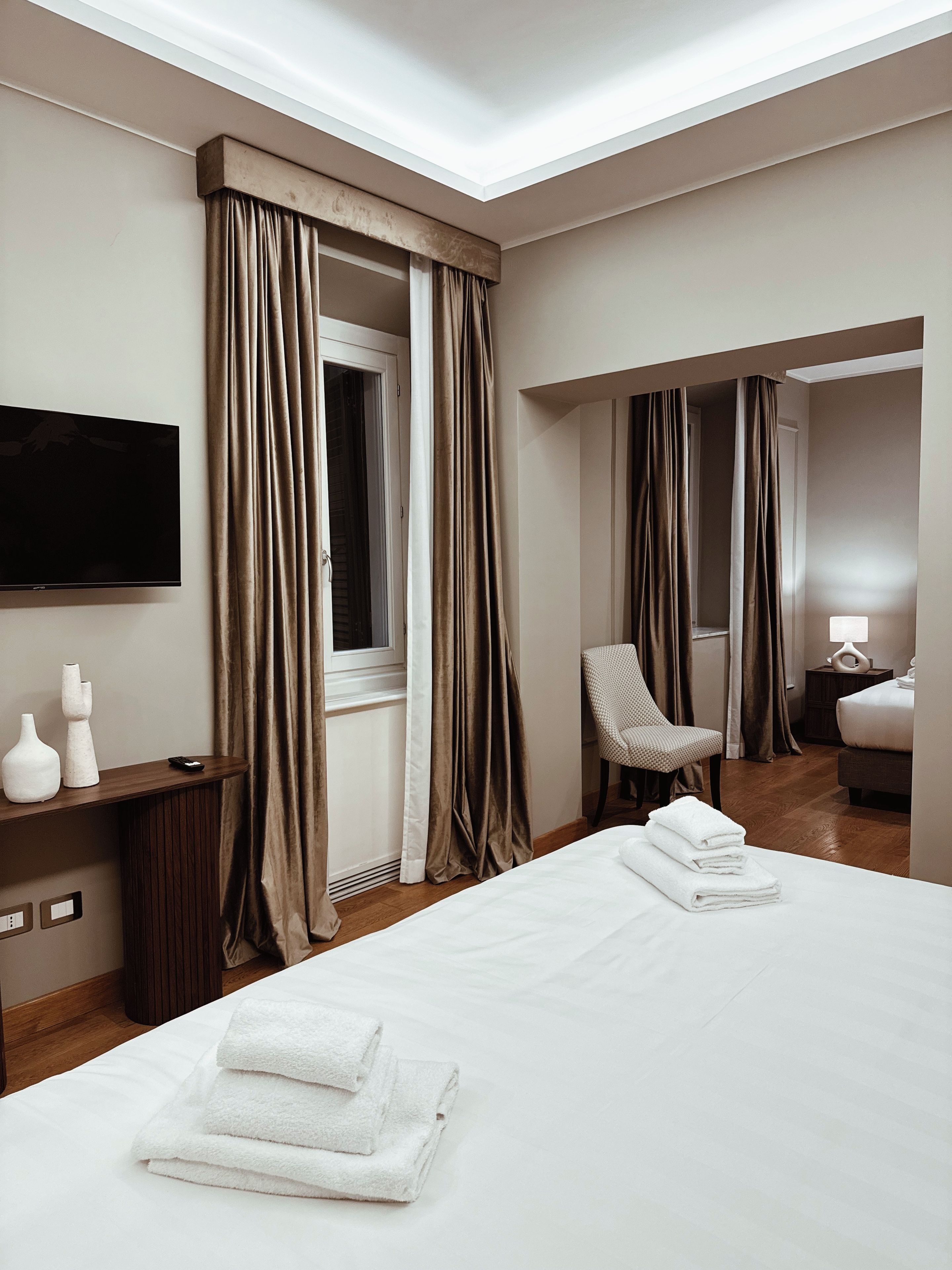 Una camera d'albergo con un letto grande, una poltrona, una scrivania con televisore a schermo piatto e un comodino con lampada.