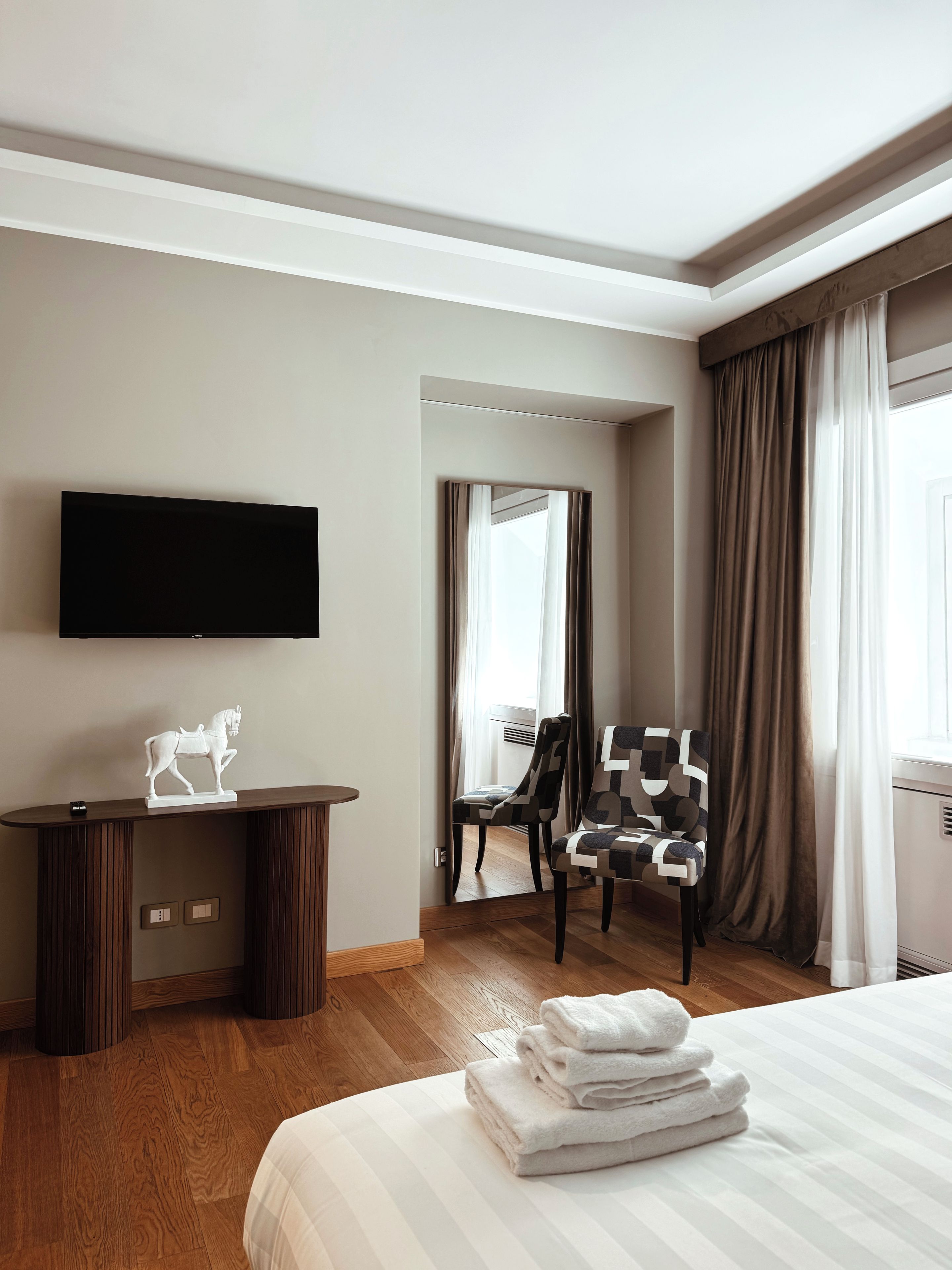 A hotel room with a bed, a flat-screen TV mounted on the wall, a desk with a statue, a chair, and a window with curtains.