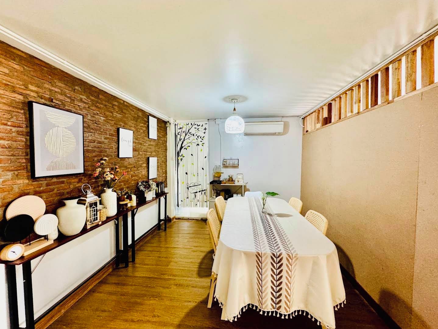 Uma sala de jantar com uma mesa comprida, cadeiras, piso de madeira, parede de tijolos e um ventilador de teto.