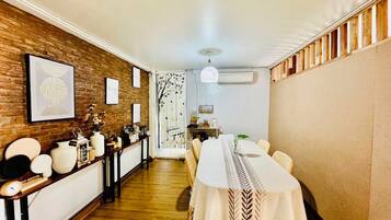 Uma sala de jantar com uma mesa comprida, cadeiras, piso de madeira, parede de tijolos e um ventilador de teto.