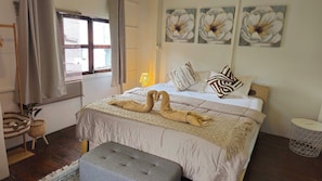A hotel room with a bed, a bedside table, a lamp, a window with curtains, and wall art. - Charming Home Chiang Mai (Chiang Mai)