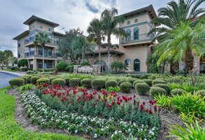 Un hôtel à plusieurs étages doté d’un jardin aménagé où l’on trouve des fleurs rouges et blanches, des palmiers et un panneau en pierre sur lequel est inscrit « Bella Piazza ».