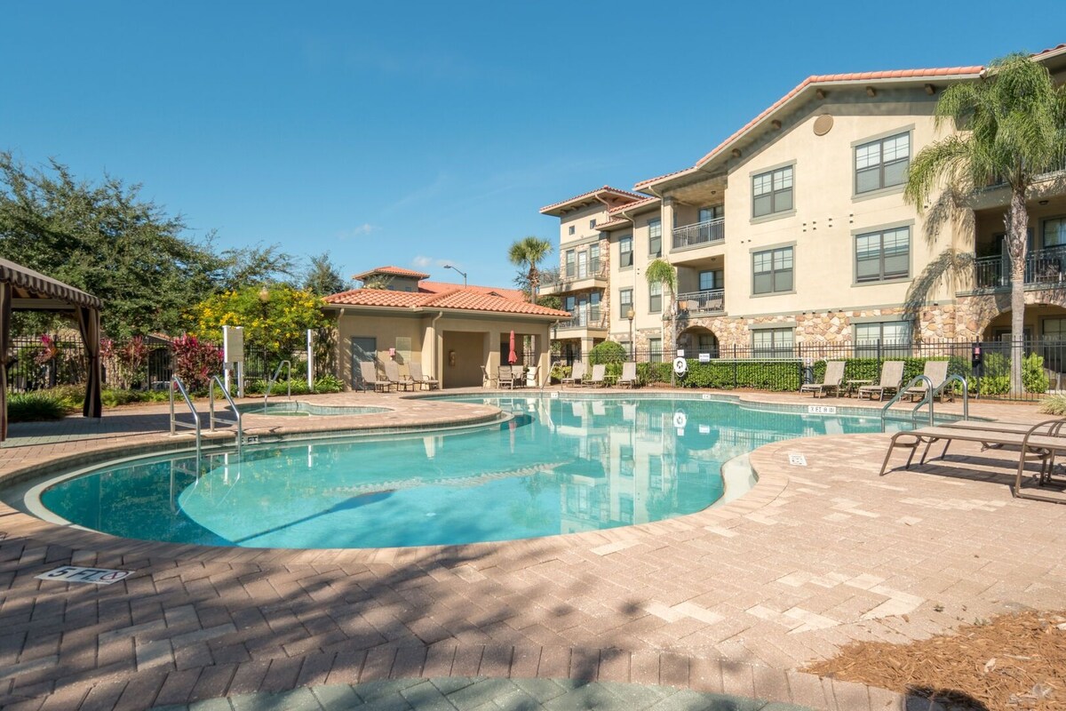 A swimming pool with lounge chairs and a covered seating area, surrounded by multi-story buildings with balconies.