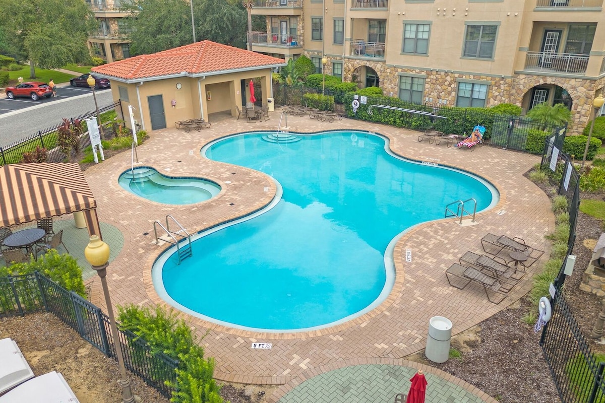A swimming pool with a unique shape, surrounded by lounge chairs and umbrellas, with a building in the background.