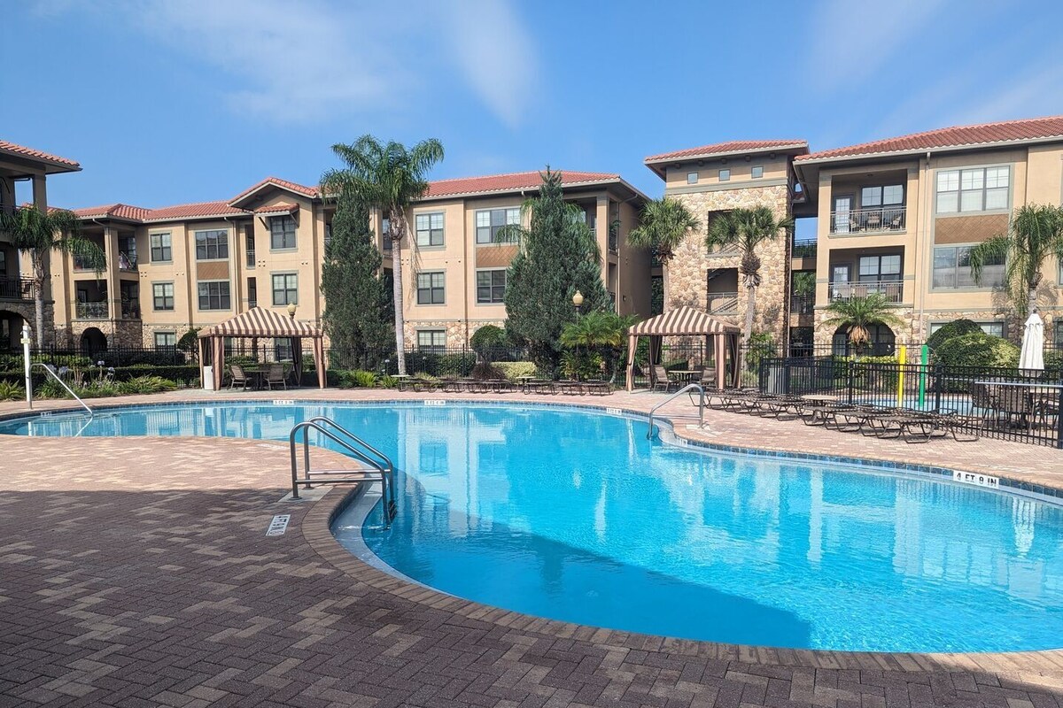 A swimming pool with a brick deck, surrounded by multi-story buildings with balconies and covered outdoor seating areas.