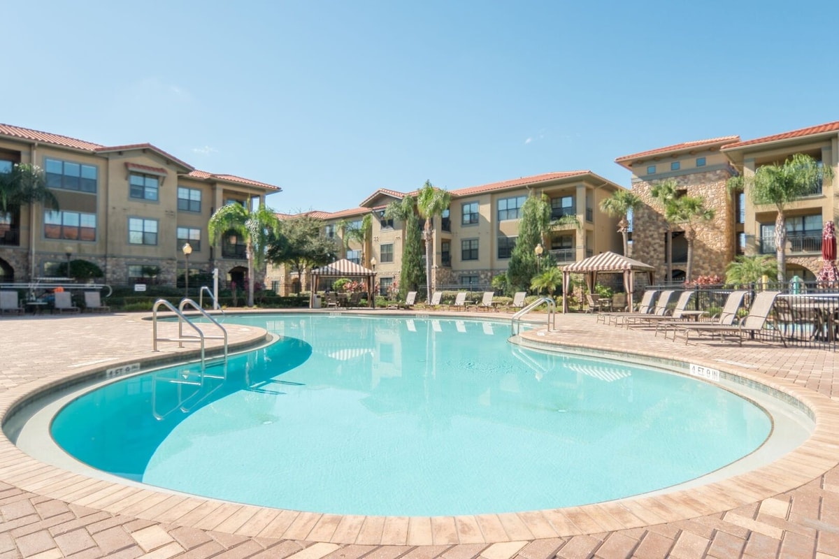 A swimming pool with lounge chairs and umbrellas, surrounded by multi-story buildings with balconies.
