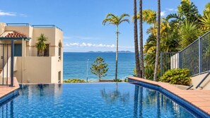 A swimming pool with a view of the ocean, palm trees, and a building with a tiled roof. - Villa Vista Three - Superb Ocean View! (Yeppoon)