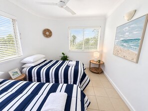 A bedroom with two beds, a ceiling fan, a window with blinds, a bedside table with a lamp, and a beach-themed painting on the wall. - Villa Vista Three - Superb Ocean View! (Yeppoon)
