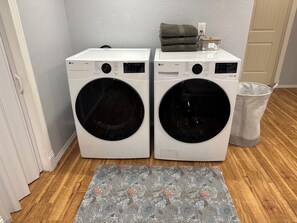 A laundry area with two white LG ThinQ front-loading washing machines, a grey rug, and a white trash can. - 2 bedroom Hillside Hideaway (Anchorage)