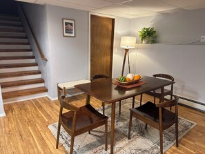 Een eetgedeelte met een houten tafel, vier stoelen, een schaal fruit, een vloerlamp en een trap op de achtergrond.