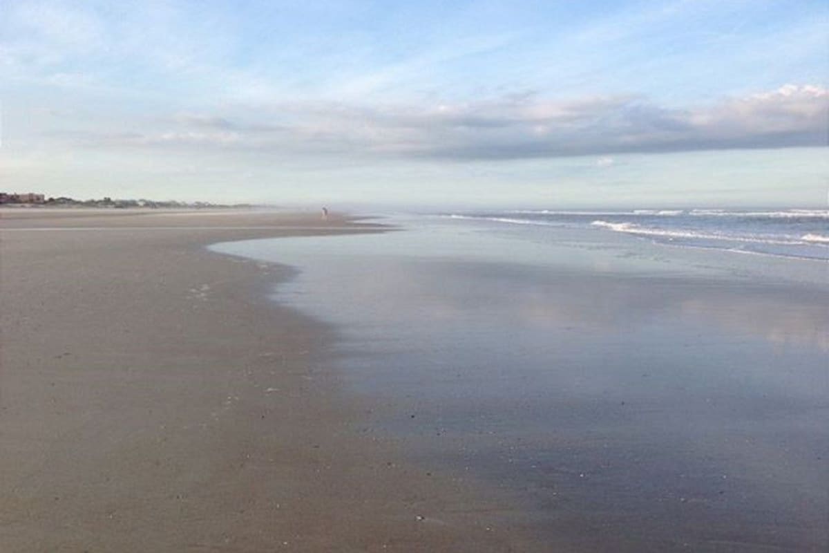 Ein weiter Sandstrand mit sanften Wellen, einem klaren Himmel und einer fernen Küstenlinie.