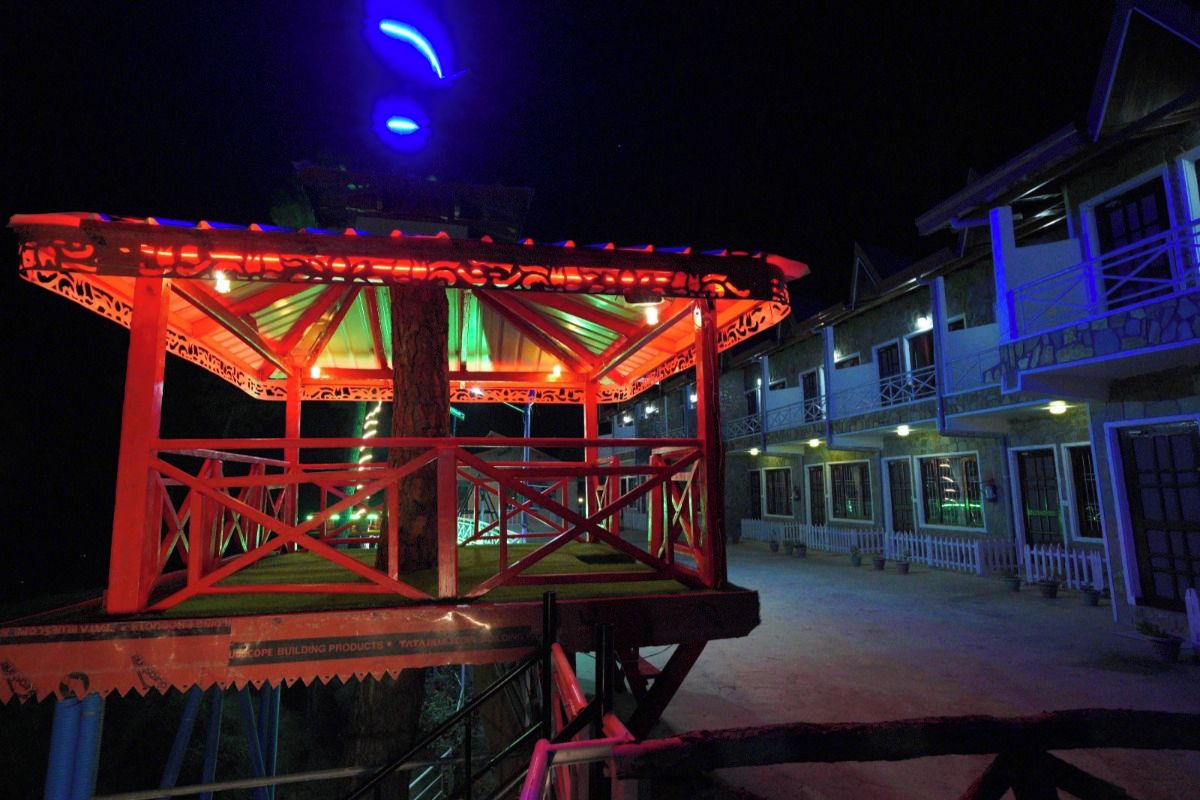 Un mirador con estructura roja y techo verde, ubicado frente a un edificio con varias ventanas y una cerca blanca.