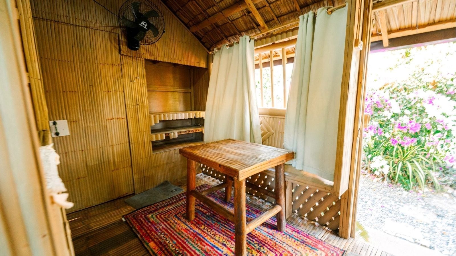 Una habitación de madera con una mesa y un ventilador, una ventana con cortinas y vistas a flores afuera.