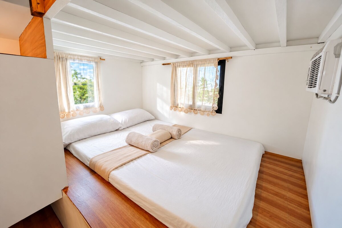 Habitación individual con cama, suelo de madera, paredes blancas y ventana con cortina.