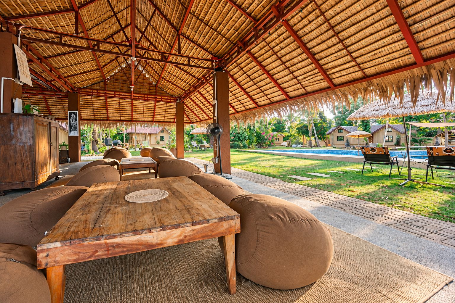 Un área de estar al aire libre con techo de paja, muebles de madera y una piscina al fondo.