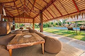 An outdoor seating area with a thatched roof, wooden furniture, and a pool in the background.