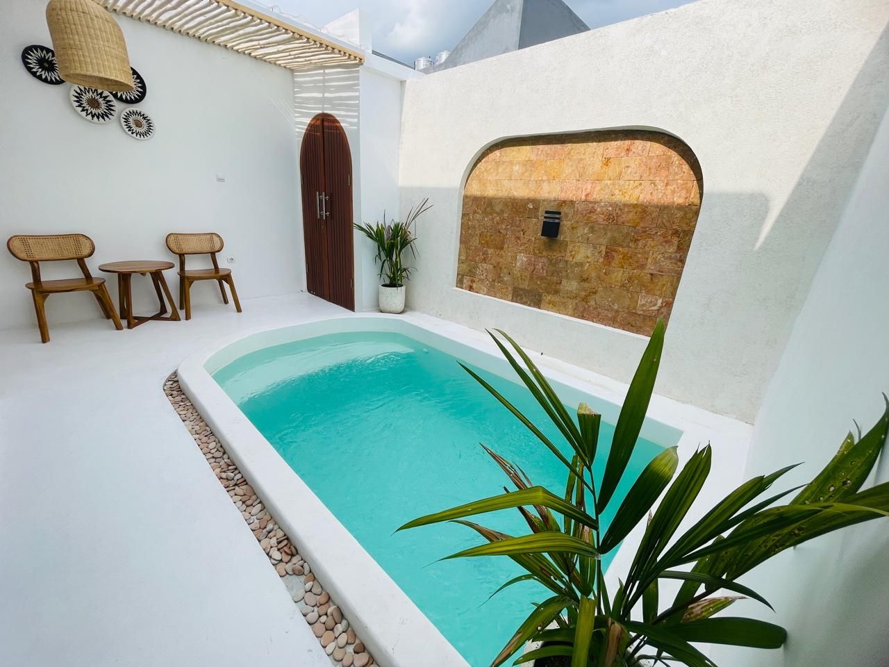 A swimming pool with a white tiled deck, surrounded by a white wall with a wooden door and a small table with chairs.