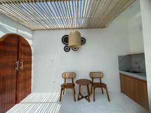 Una habitación con puerta de madera, techo tejido, lámpara de pared y una pequeña cocina con lavamanos.