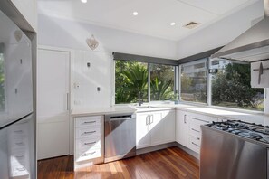 A modern kitchen with white cabinetry, a dishwasher, a stove, and large windows offering a view of greenery. - Verney Beachside Getaway Two (Kings Beach)
